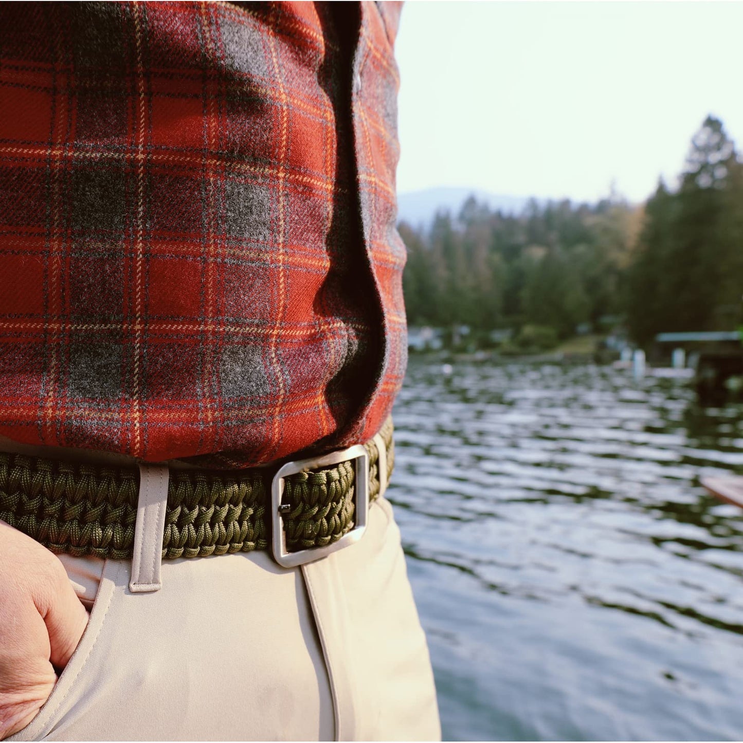 Person wearing a red plaid shirt and beige pants with a green belt, standing by a lake with trees in the background.