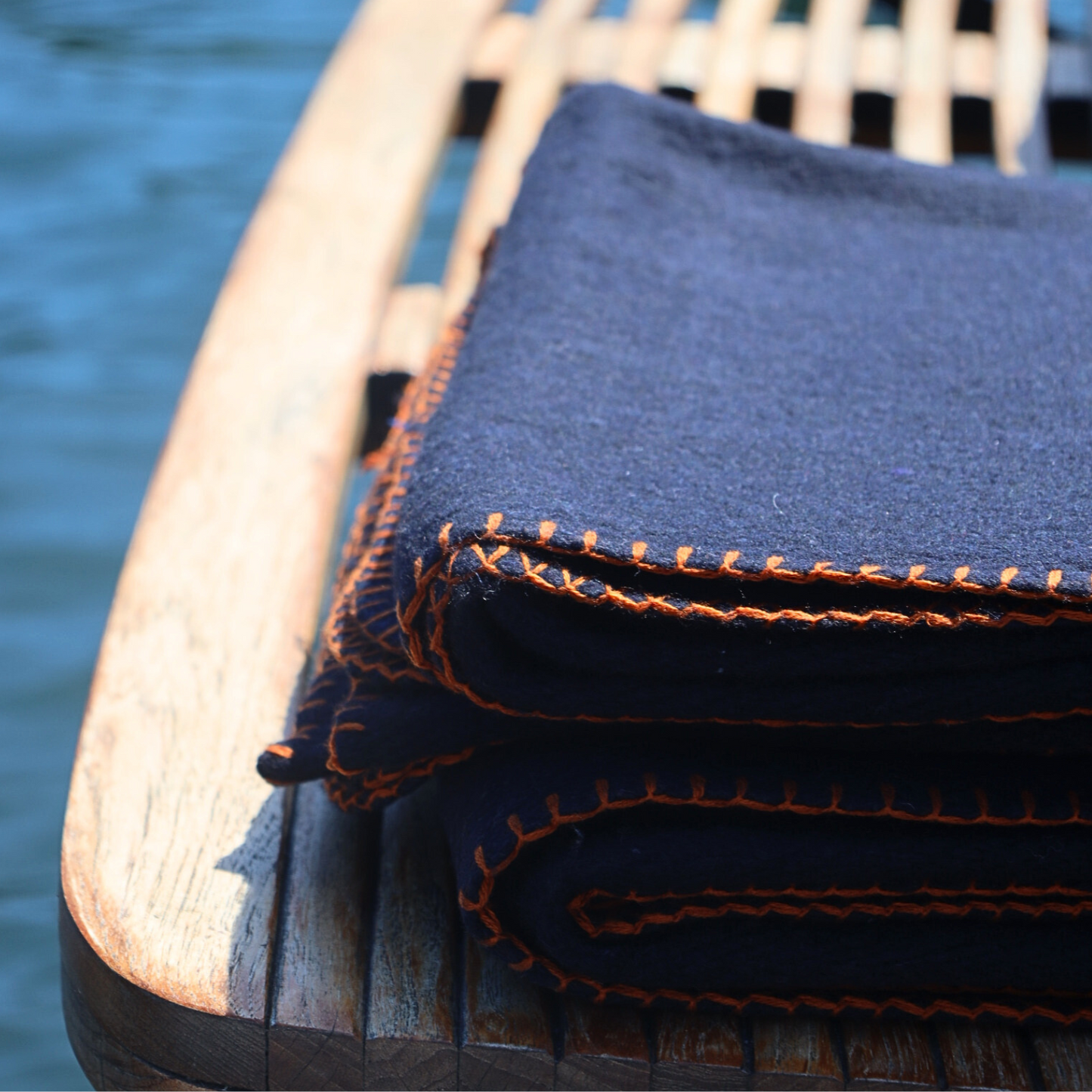 Folded blue blanket with orange stitching on a wooden chair by a body of water.