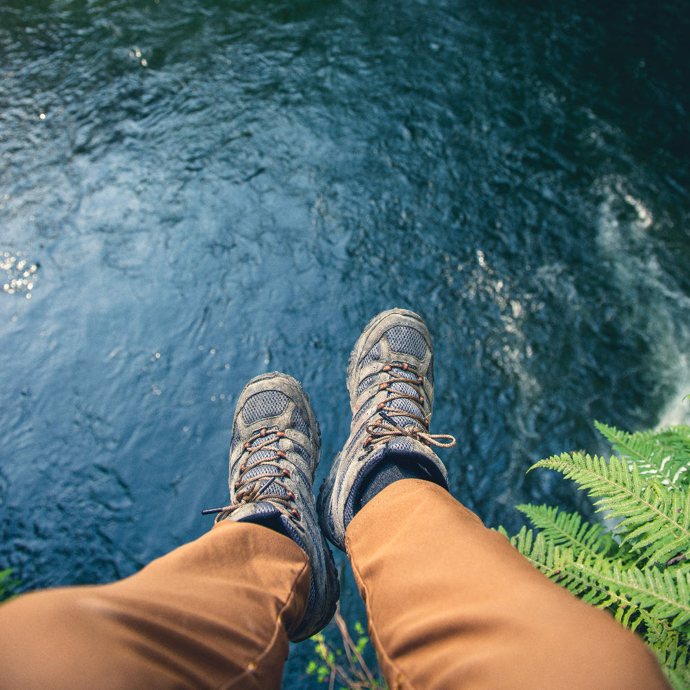 Person wearing hiking boots standing on a rock above a river with greenery around