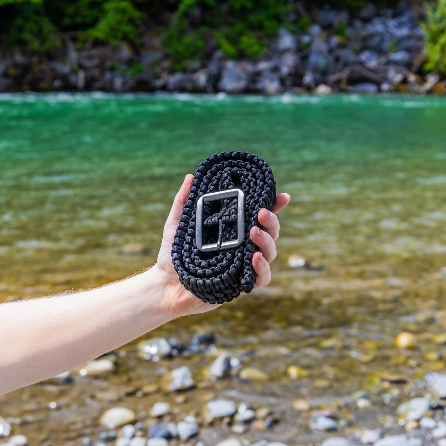 Person holding a black paracord belt with a buckle against a natural background of water and rocks.