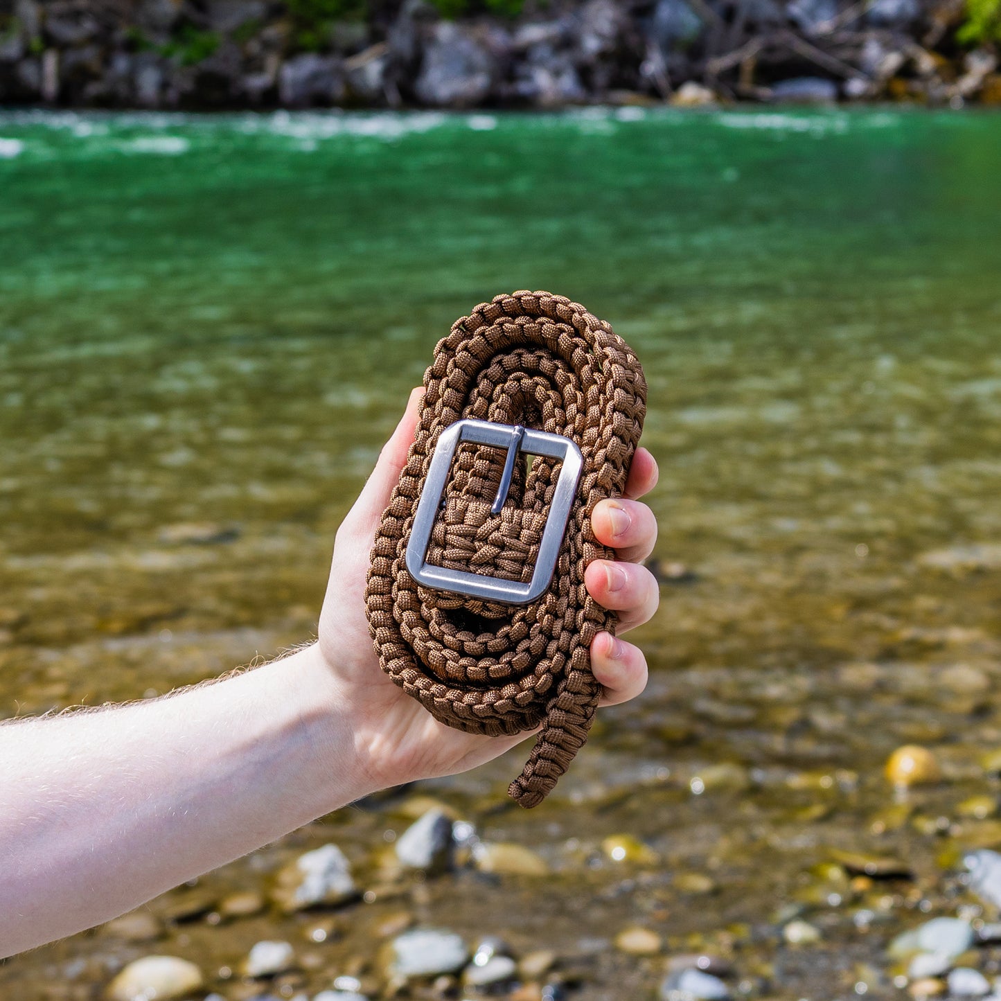 Hand holding a brown woven belt with a silver buckle against a natural background of water and rocks.